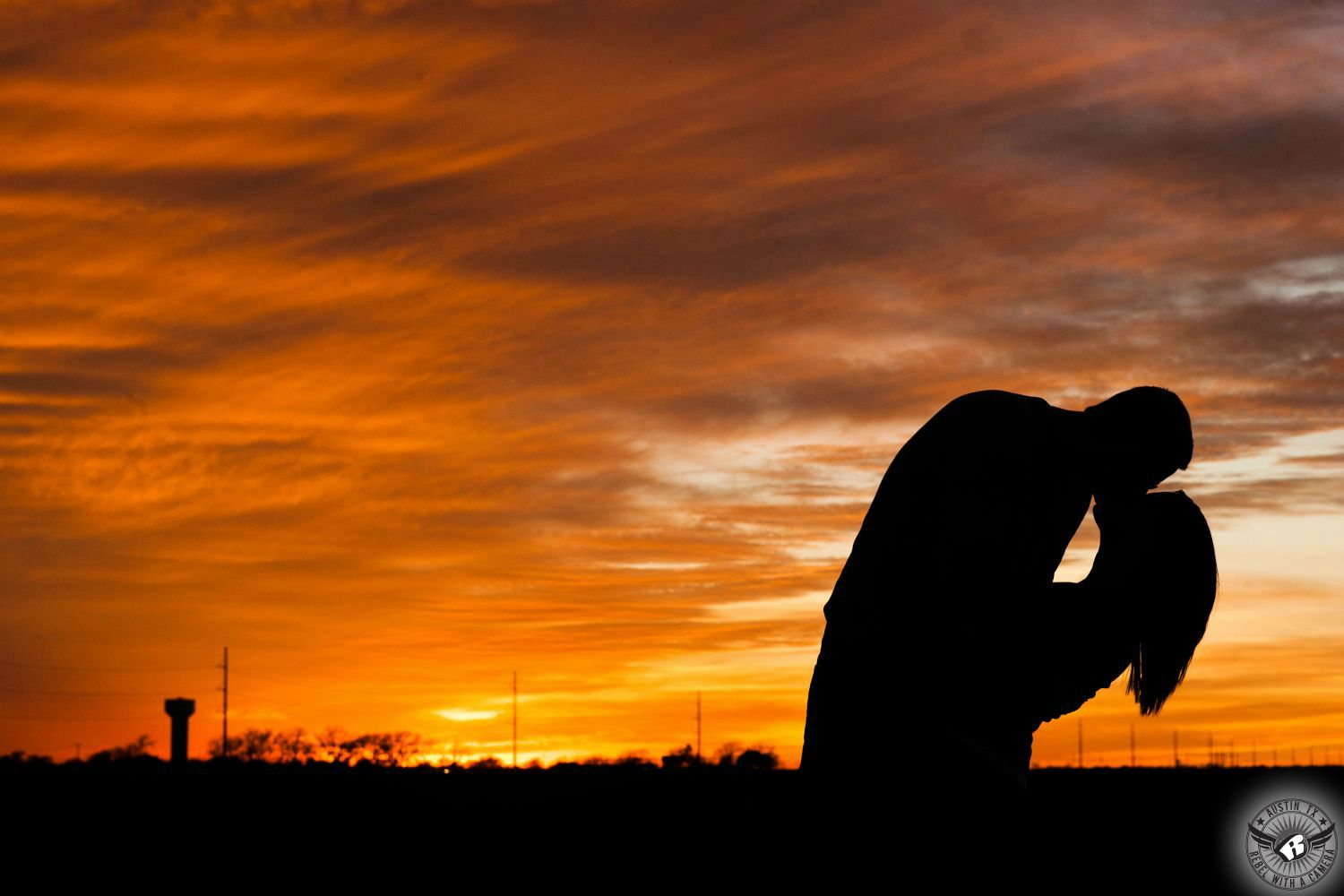 A silhouette of a man and a woman embrace and kiss while dipping with the woman's hair dangling below with a big orange cloudy sky in the background with a water tower silhouette in the distance in this breathtaking engagement portrait in Taylor in rural Austin, Texas.